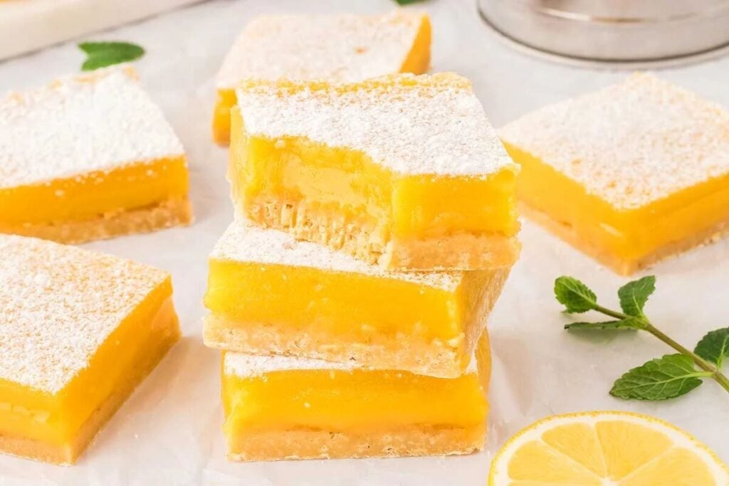 Stack of gluten free lemon bars dusted with powdered sugar, garnished with mint leaves and lemon slices on a marble surface.