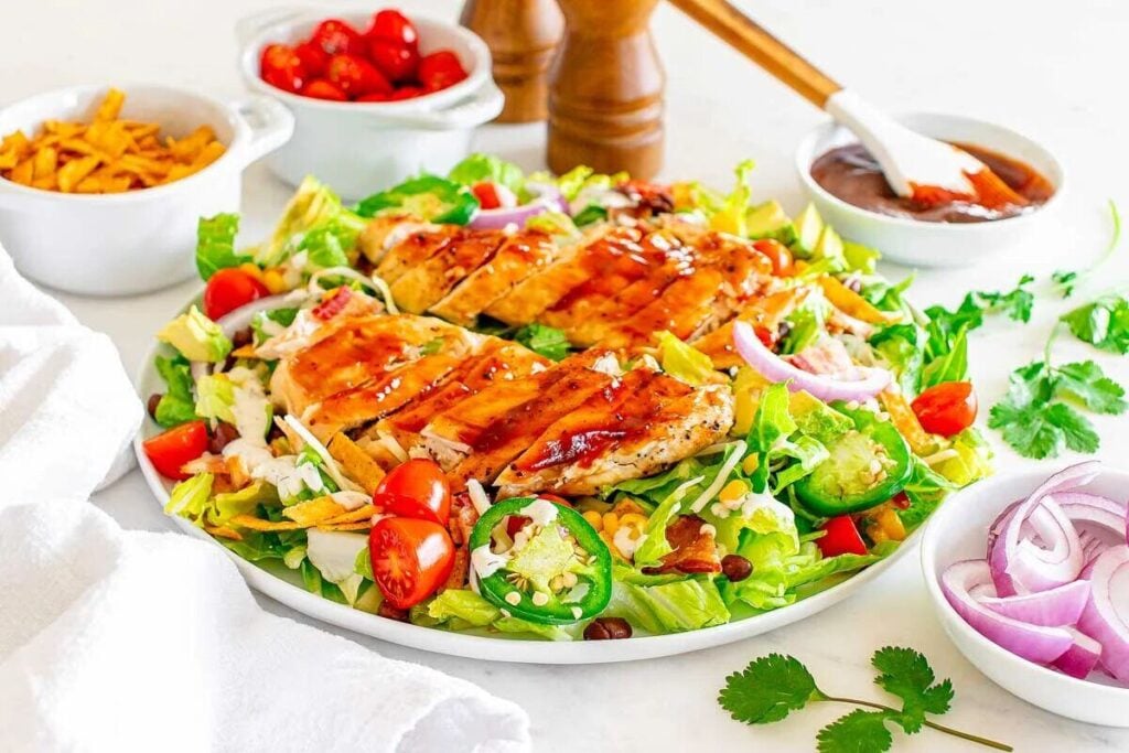 A grilled BBQ chicken salad with lettuce, cherry tomatoes, red onion, corn, and tortilla strips is arranged on a plate with dressing, condiments, and seasonings in the background.
