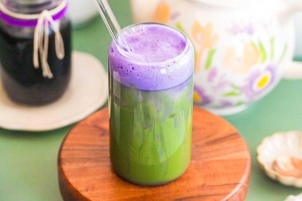 A glass with an iced lavender matcha latte topped with purple foam sits on a wooden coaster, accompanied by a floral teapot and a small shell.