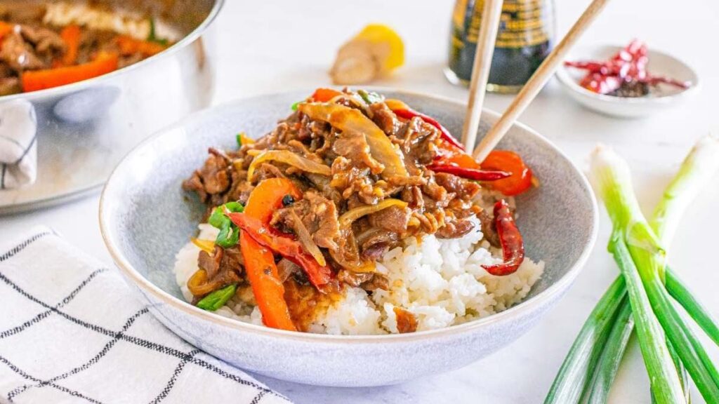 A bowl of rice topped with stir-fried beef and vegetables, with chopsticks. Green onions, a pot, and condiments are in the background.
