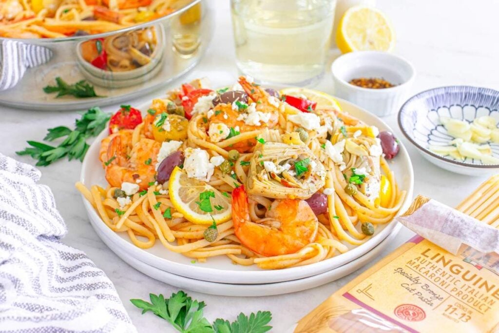A dish of Mediterranean shrimp linguine topped with vegetables, herbs, and feta cheese is served on a white plate. 
