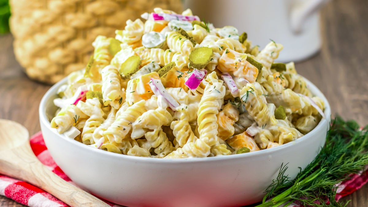 Bowl of pasta salad with rotini, diced red onion, pickles, dill, and creamy dressing, placed on a red-checked cloth.