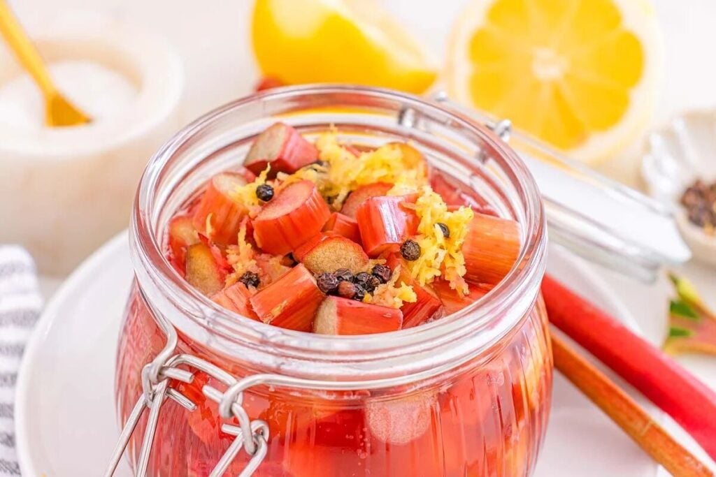 Glass jar filled with pickled rhubarb, lemon zest, and spices, with fresh lemons and rhubarb in the background.