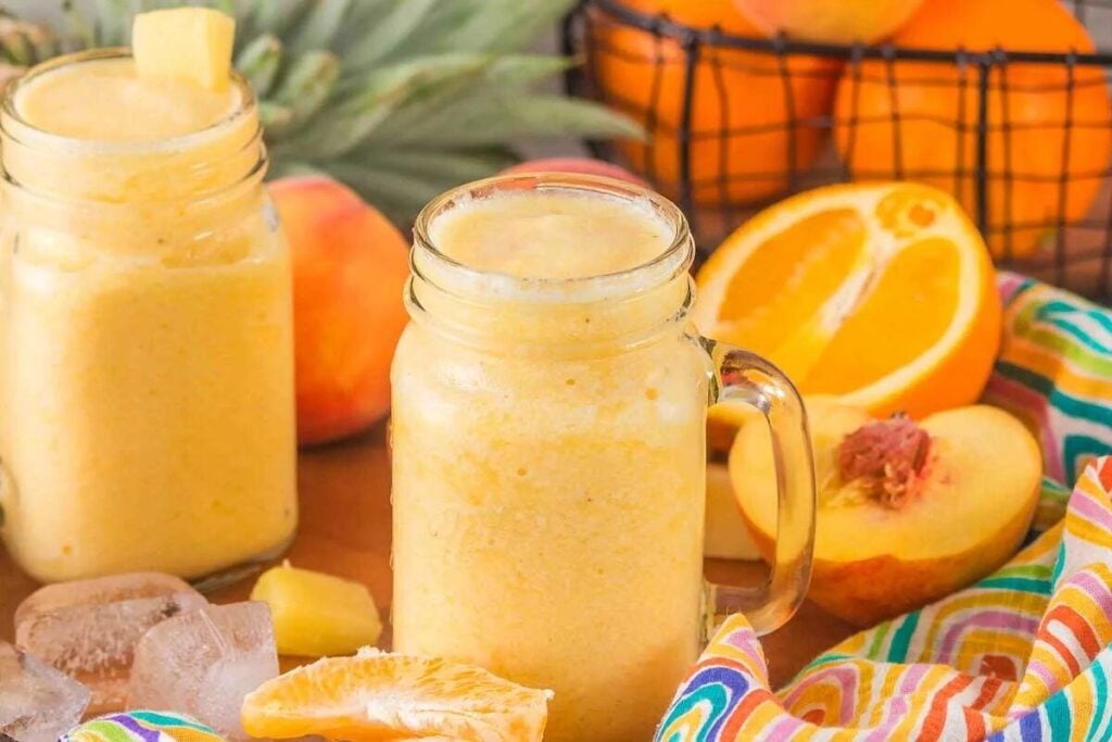 Two mason jars of Pineapple Peach Smoothie with fruit slices, ice cubes, and a colorful cloth nearby.