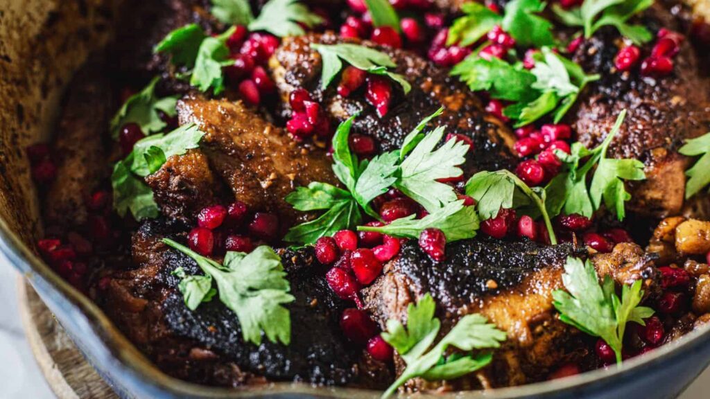 A close-up of a cooked dish featuring several pieces of meat topped with vibrant green parsley leaves and scattered pomegranate seeds. The dish is placed in a dark pot.