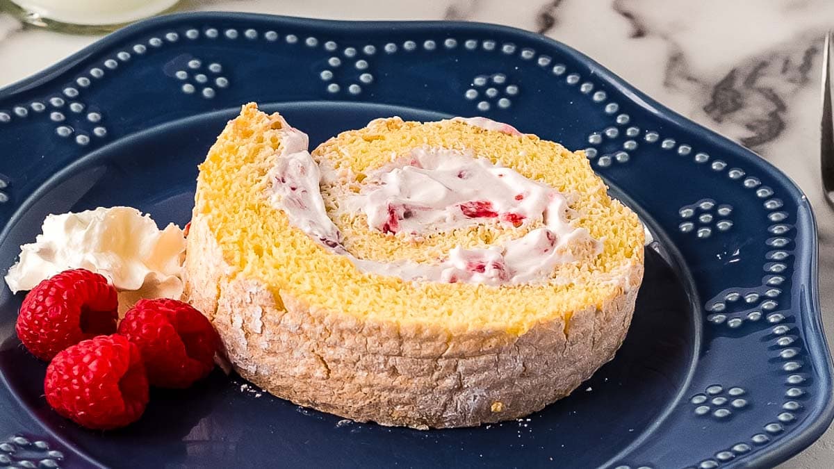 A slice of swiss roll cake with cream and raspberries on a blue plate, garnished with dollop of whipped cream and three raspberries.