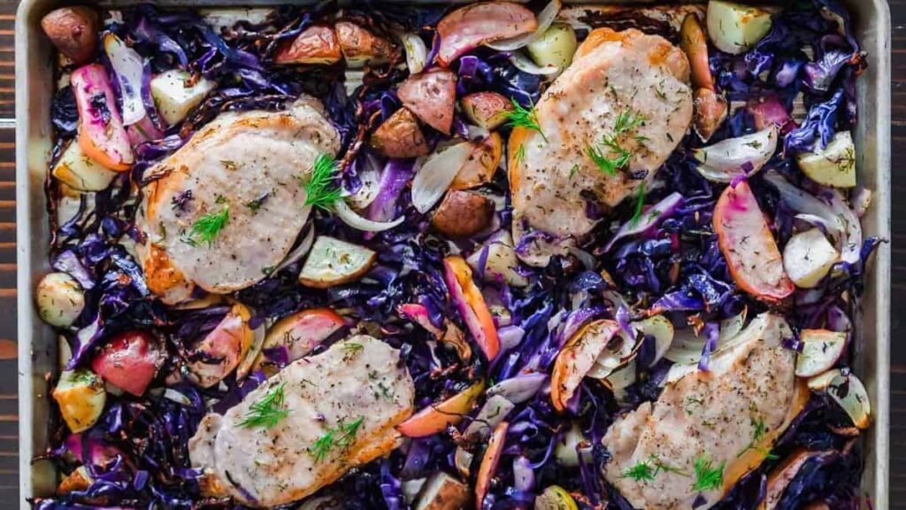A baking sheet with four pork chops surrounded by red cabbage, potatoes, and sliced apples.