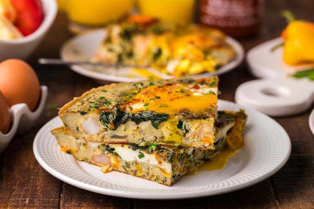 Two slices of vegetable frittata on a white plate, resembling a seafood omelette, with eggs and fresh vegetables in the background.