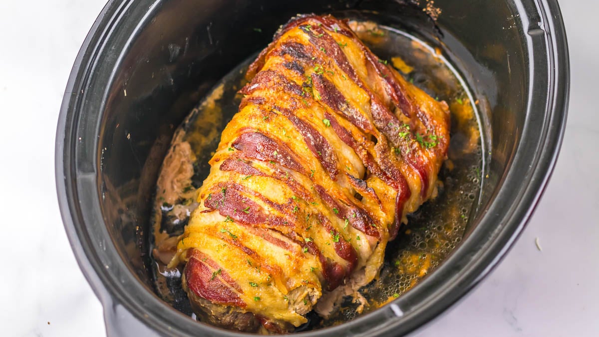 Bacon-wrapped pork loin cooked in a slow cooker, with juices at the bottom and garnished with herbs.
