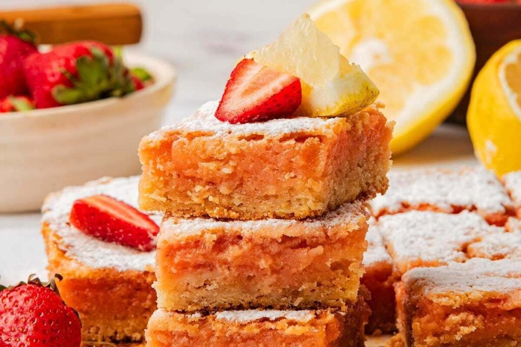 A tempting stack of strawberry lemonade bars topped with fresh strawberries and lemon slices, all dusted with a light sprinkle of powdered sugar.