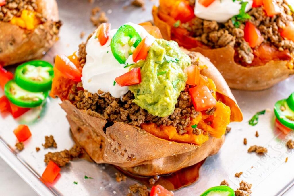 Loaded taco stuffed sweet potato topped with spiced ground meat, guacamole, sour cream, diced tomato, and sliced jalapeños on a tray.