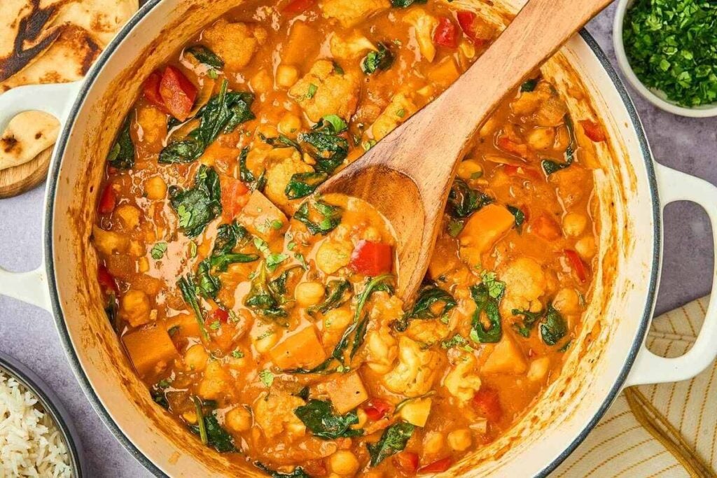 A pot of fragrant vegetable curry brimming with chickpeas, spinach, and cauliflower is gently stirred with a wooden spoon. Nearby, rice and naan await alongside a small bowl of fresh herbs.