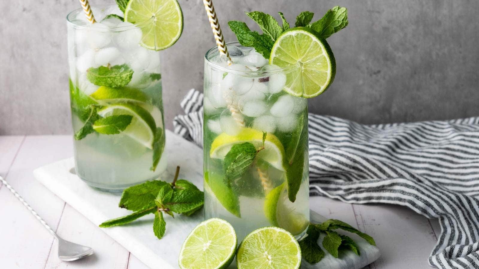 Two glasses of mojito with ice, lime slices, and mint leaves, each with a striped straw, placed on a white board with extra lime and mint nearby.