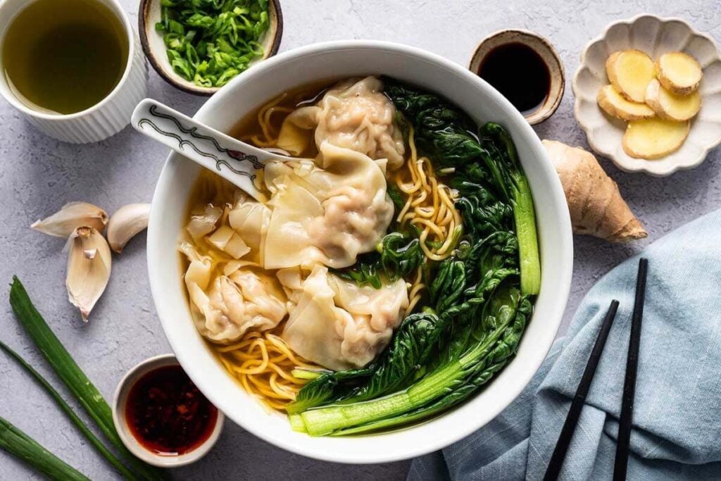 A steaming bowl of wonton noodle soup with vibrant greens is surrounded by small bowls of condiments, garlic, and ginger slices, all perfectly arranged on a sleek gray surface.