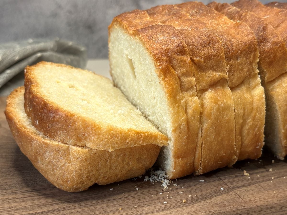 A sliced loaf of white bread rests on a wooden surface, revealing its light, fluffy interior and golden crust.