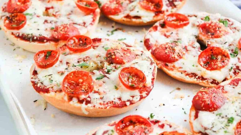 Bagels topped with melted cheese, tomato sauce, and pepperoni, garnished with herbs on a baking tray.