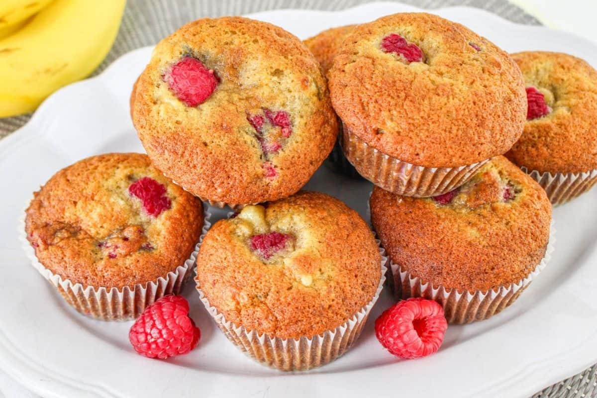 A plate of banana raspberry muffins with raspberries on top and bananas in the background.