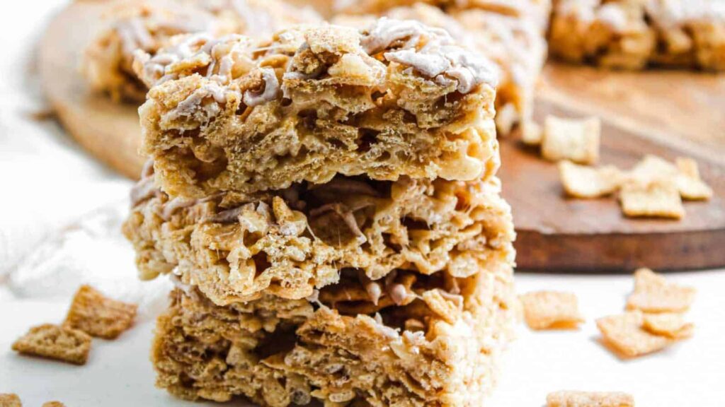 Three stacked cereal bars made with cinnamon cereal and drizzled with icing, with loose cereal pieces around.