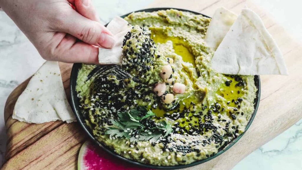 A bowl of green hummus garnished with black sesame seeds, chickpeas, and herbs sits on a wooden board. A hand dips a piece of pita bread into the hummus. Slices of pink and white radish are visible on the board.