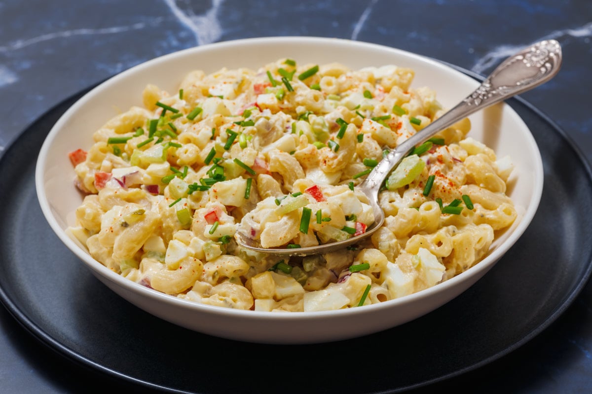 A bowl of Deviled Egg Pasta Salad with chopped vegetables and herbs, is set on a black plate, and a spoon rests in it.