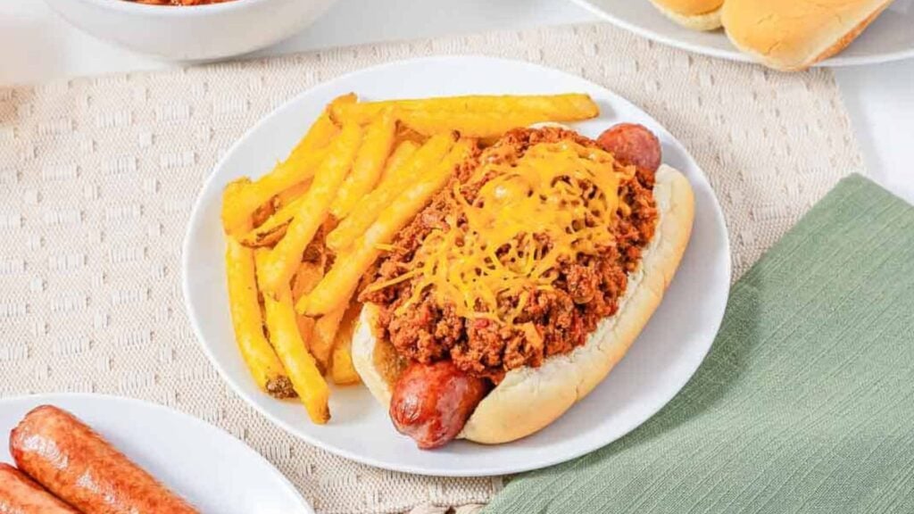 Chili cheese hot dog with French fries on a white plate, set on a beige placemat next to a green napkin.