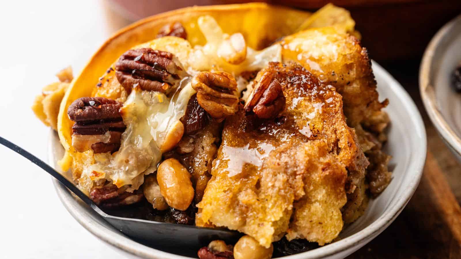A bowl of bread pudding topped with pecans, walnuts, and a drizzle of syrup, served with a spoon.