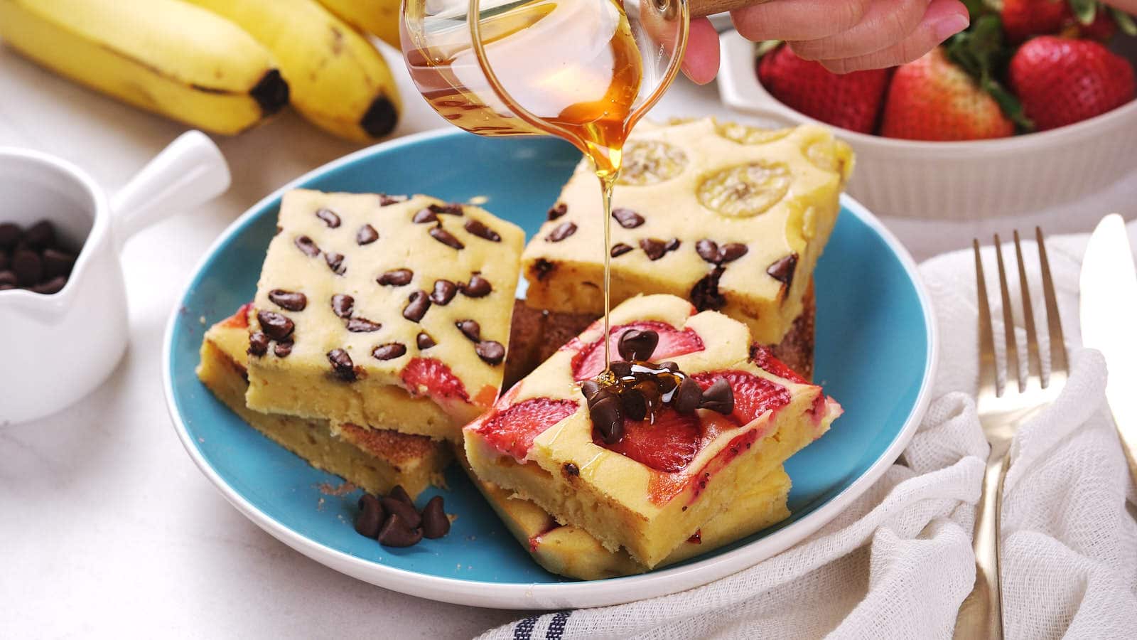 Three pieces of sheet pan pancakes topped with banana, strawberries, and chocolate chips on a blue plate.
